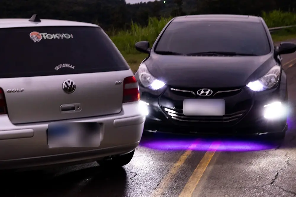 Hyundai Elantra preto com farol de LED aceso e neon roxo embaixo do carro, ao lado de um Golf com adesivo da Tokyo Car