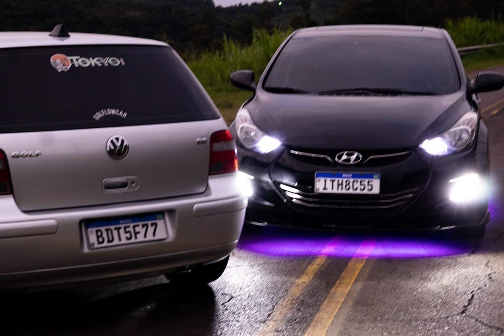 Hyundai Elantra preto com farol de LED aceso e neon roxo embaixo do carro, ao lado de um Golf com adesivo da Tokyo Car